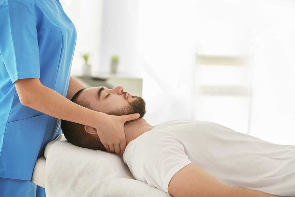 Adult performing guided back exercises on a treatment table beside a clinician in a bright rehabilitation room.